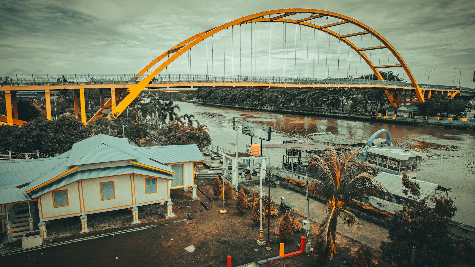 Jembatan lengkung oranye di atas sungai dengan rumah panggung di tepinya